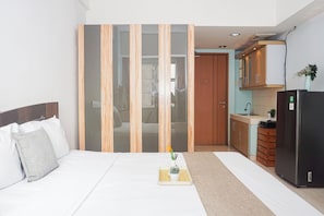 A bedroom with a bed, a bedside table with a vase and a plant, and a view into a kitchen area with cabinets and a refrigerator. - Compact Minimalist Studio At Apartment Margonda Residence 3 (Depok)