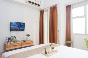 Habitación de hotel con cama, televisor montado en la pared, cómoda con libros y una planta, y ventana con cortinas.