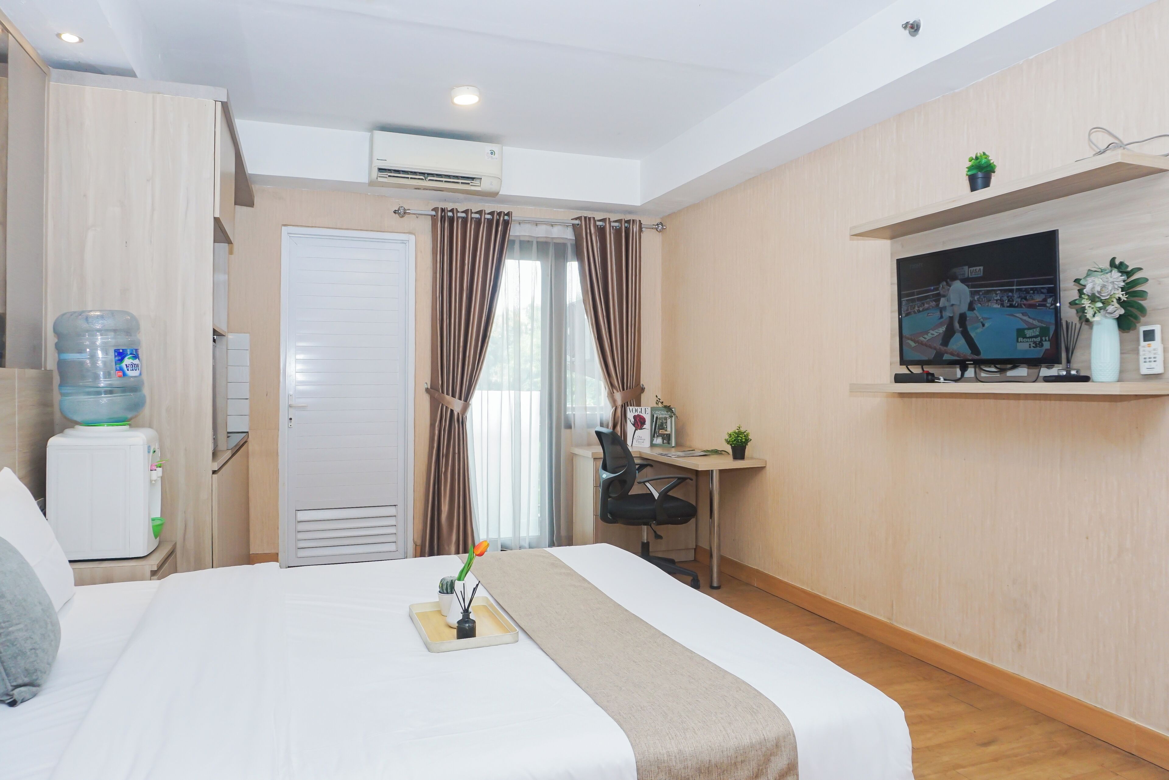 A hotel room with a bed, a desk, a TV, a water dispenser, and a window with curtains.
