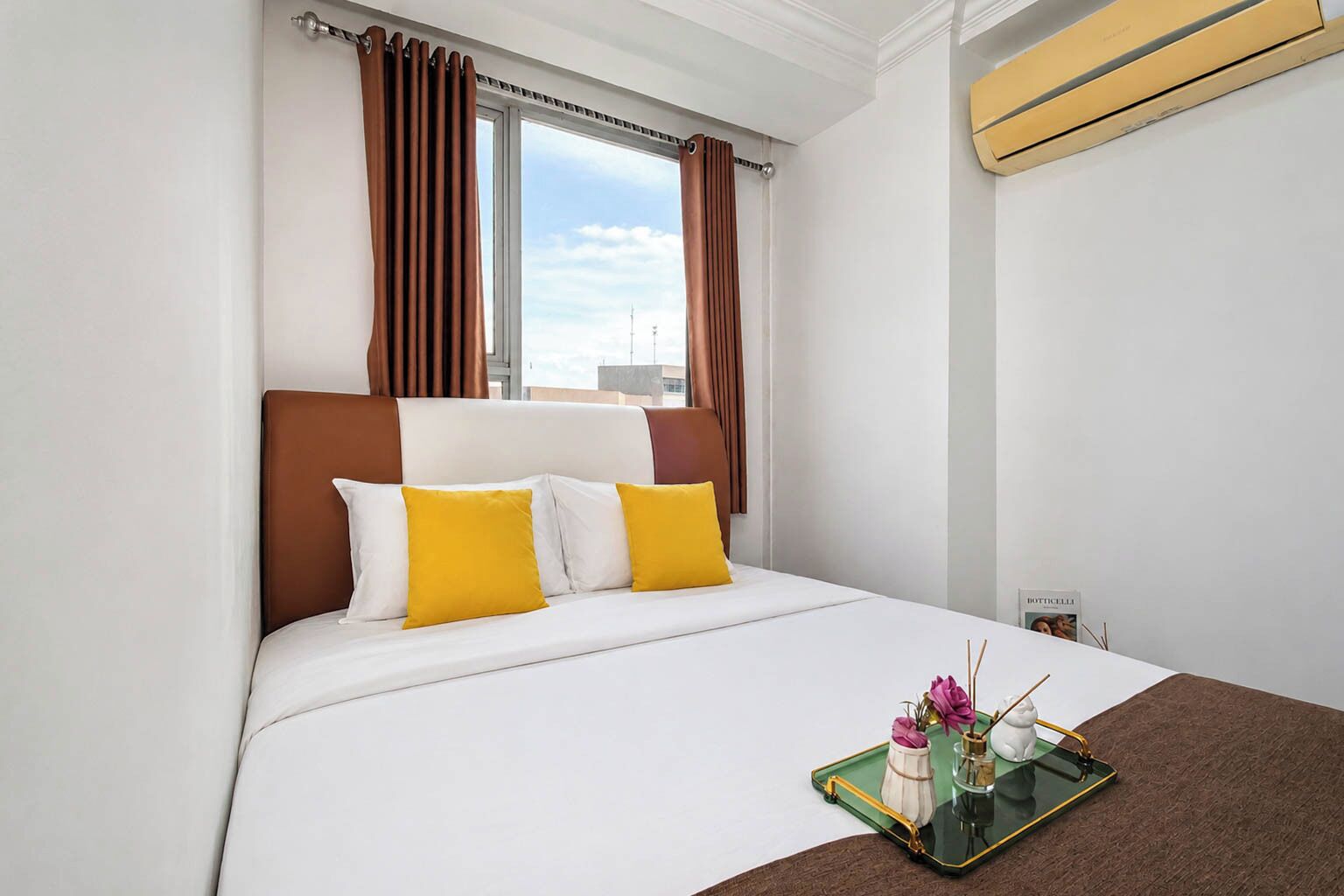 A hotel room with a bed, brown curtains, a window, and a tray with a vase and candle.