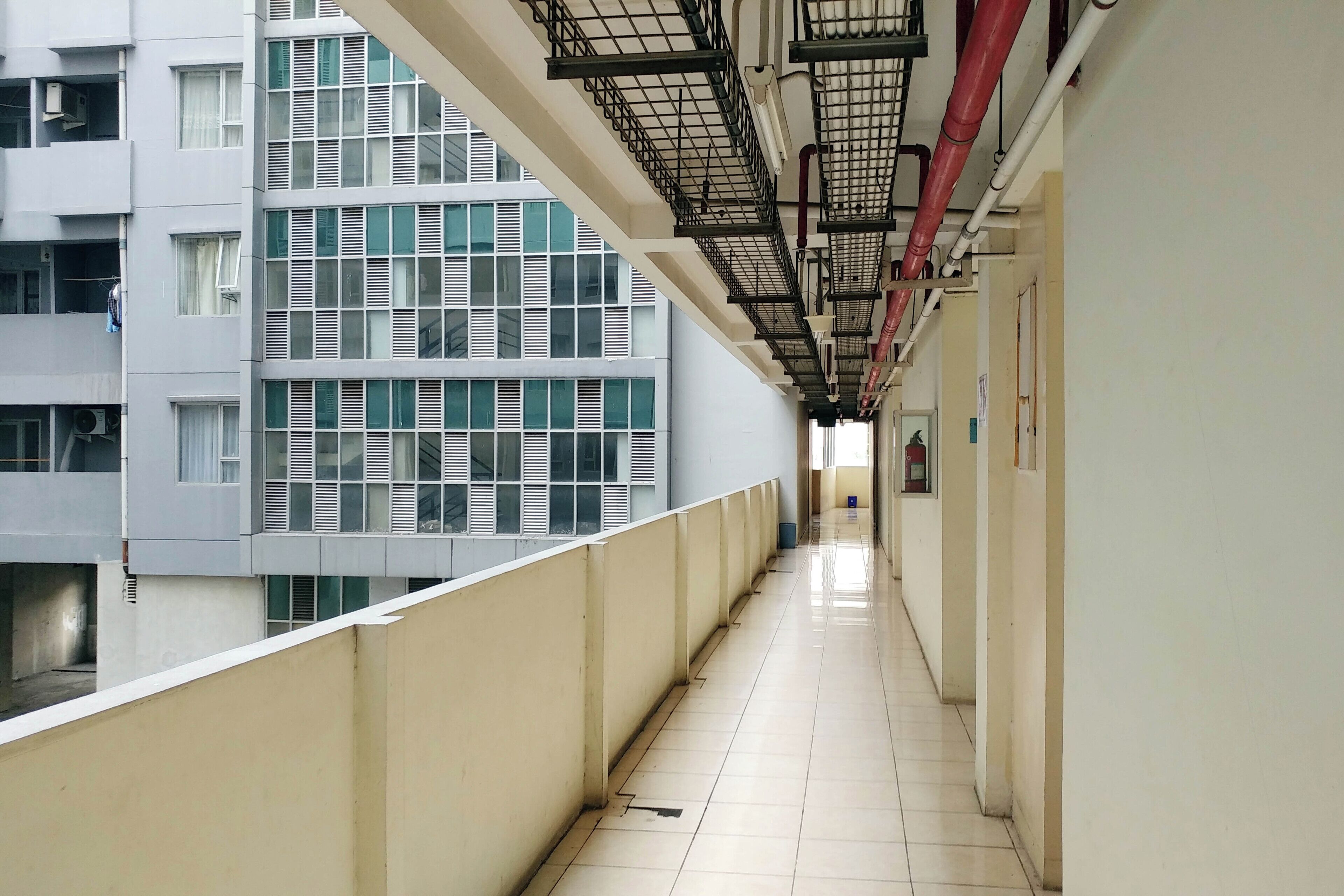 A corridor with a tiled floor, a metal railing on one side, and a view of a building with multiple windows and air conditioning units.