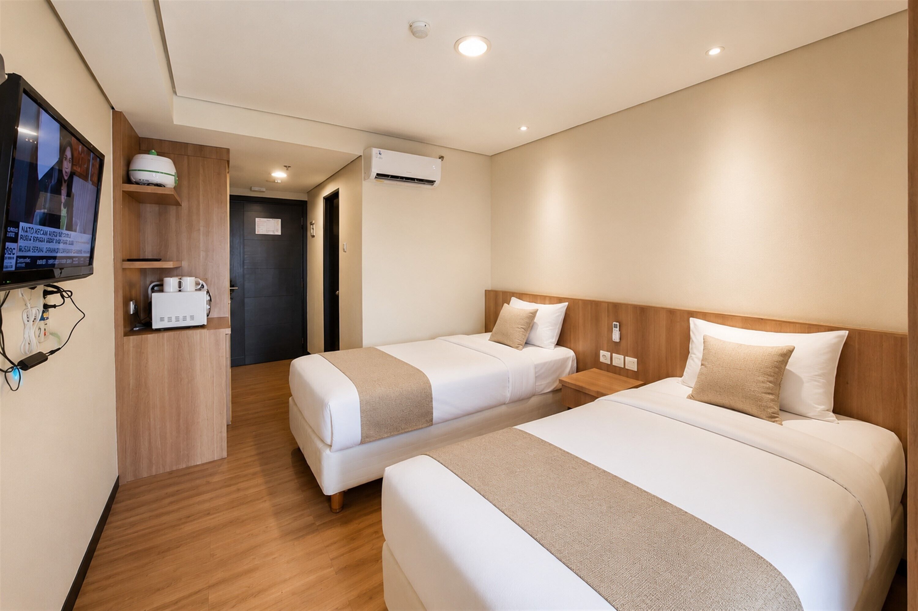 A hotel room with two beds, a TV, a desk with a computer, and an air conditioning unit.