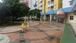 Un área exterior de ejercicios con equipo de fitness, rodeada por un patio con suelo de baldosas y follaje.