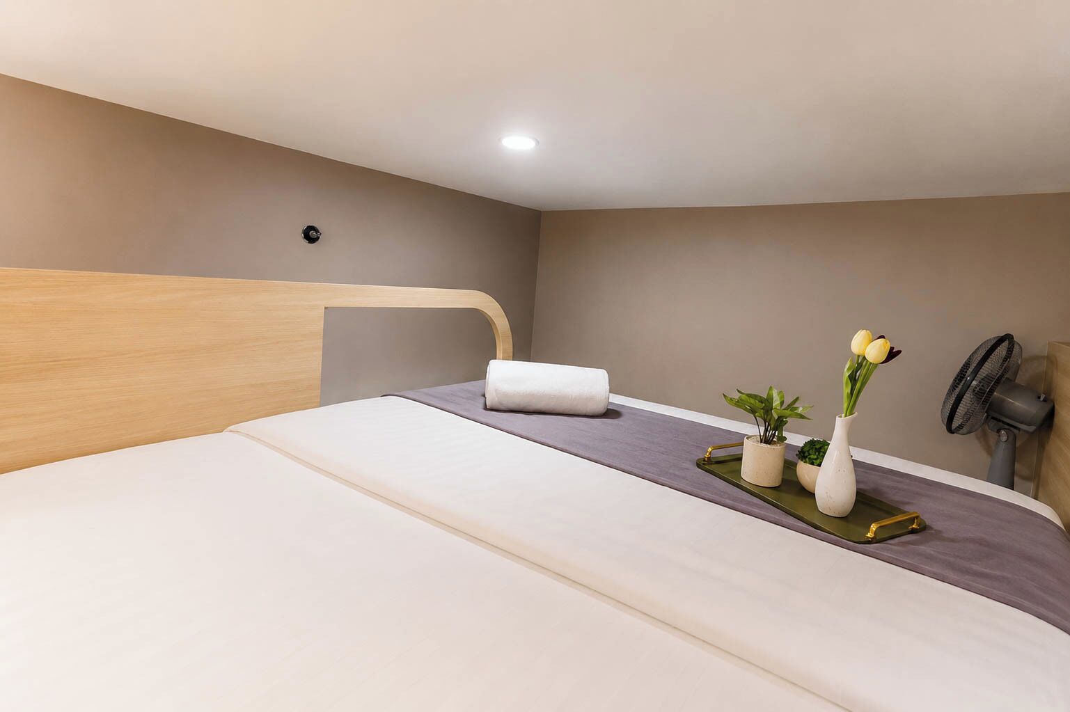 Ein Hotelzimmer mit Bett, Nachttisch, Wandventilator und einer kleinen Topfpflanze.