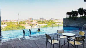 An outdoor swimming pool with a view of a cityscape, surrounded by lounge chairs and a wooden deck. - Urban Minimalist Studio At Apartment Tamansari Papilio (Surabaya)