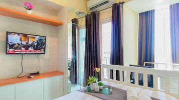 A hotel room with a bed, a TV mounted on the wall, a window with curtains, and a small plant on the bedside table.