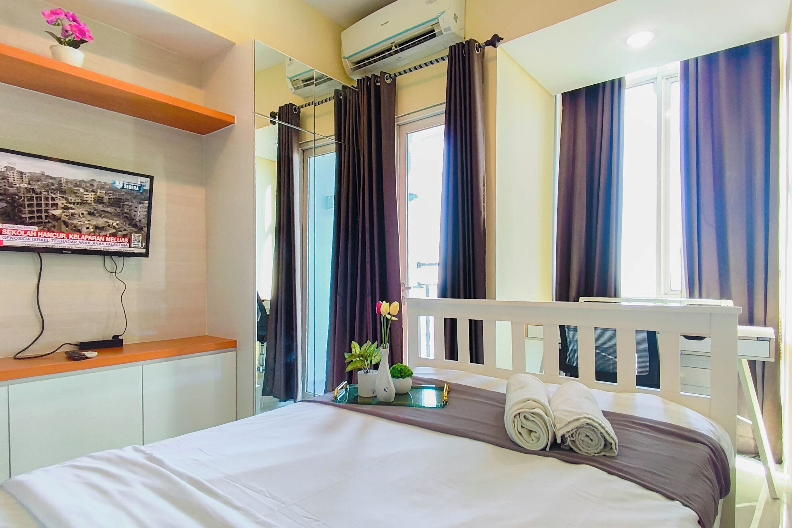 A hotel room with a bed, a television mounted on the wall, a window with curtains, and a small table with a vase of flowers.