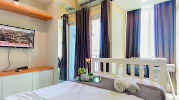 A hotel room with a bed, a television mounted on the wall, a window with curtains, and a small table with a vase of flowers.