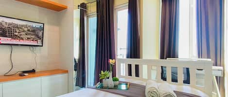 A hotel room with a bed, a television mounted on the wall, a window with curtains, and a small table with a vase of flowers.