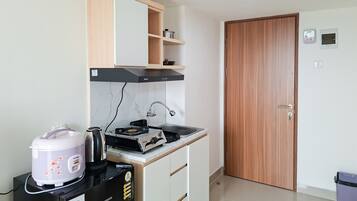 Cocina compacta con estufa, hervidor y cocedor de arroz. Hay una puerta de madera y un pequeño estante sobre la estufa.