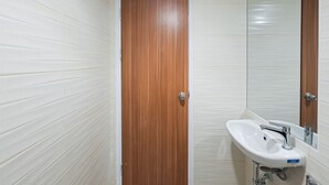 A bathroom with white tiled walls, a wooden door, and a white sink with a mirror above it. - Comfortable Nice Studio Room At Apartment Cisauk Point (Cisauk)
