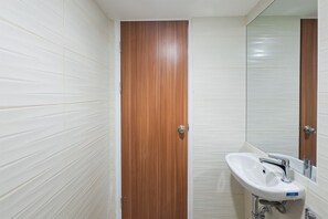 A bathroom with white tiled walls, a wooden door, and a white sink with a mirror above it. - Comfortable Nice Studio Room At Apartment Cisauk Point (Cisauk)