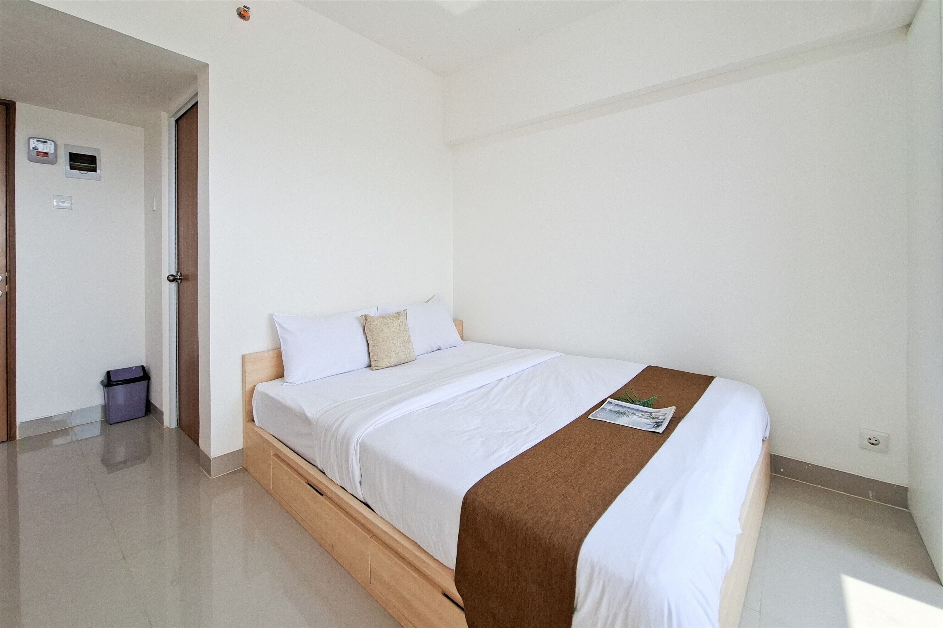 Quarto de hotel com uma cama de solteiro, cabeceira de madeira, parede branca, uma porta, uma lixeira e uma pequena mesa com uma revista.