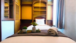 A hotel room with a bed, a small table with a plant, a folded towel, and a shelf with a plant.