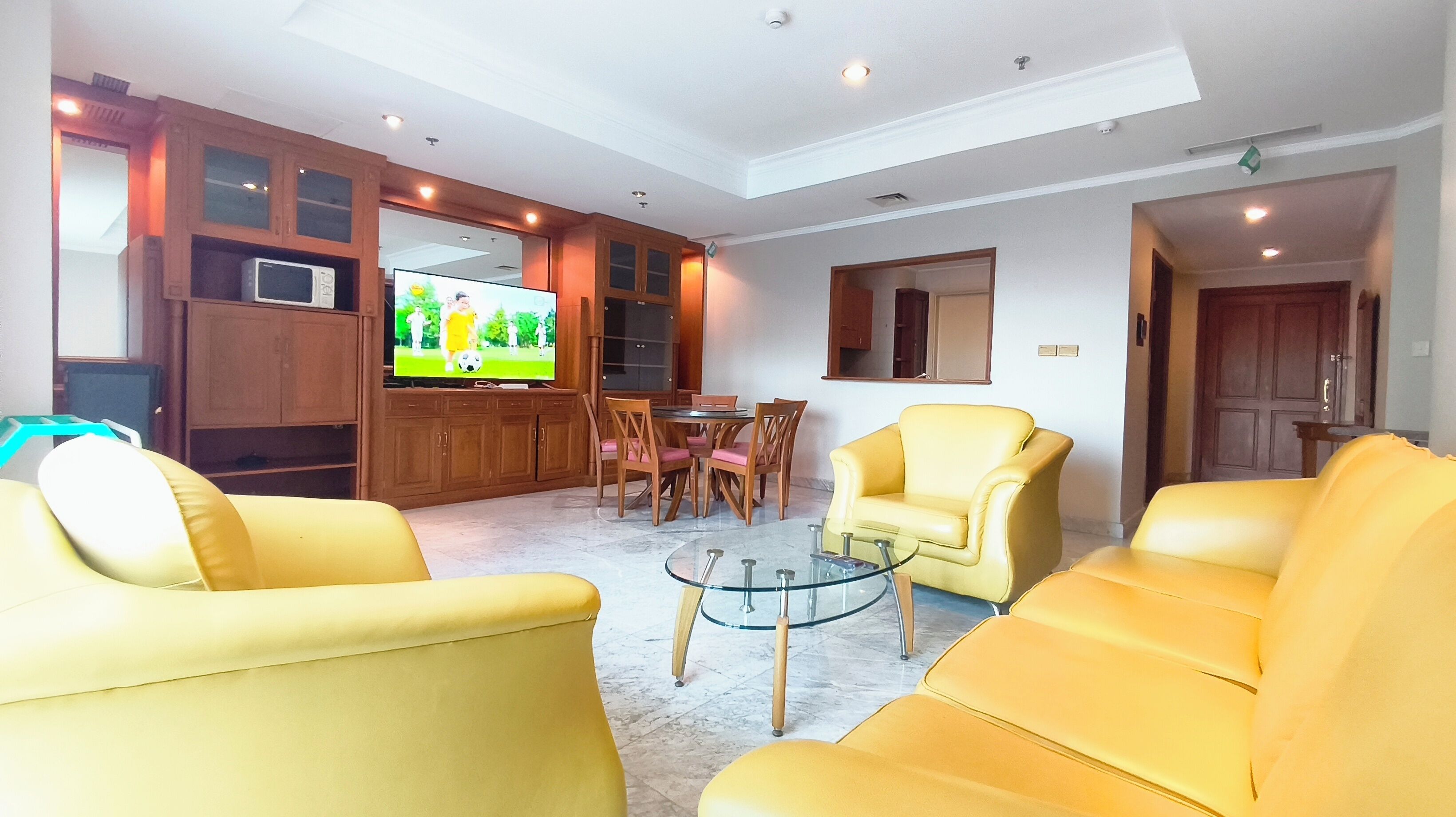 A living room with yellow sofas, a glass coffee table, wooden cabinets, a television, and a dining area with chairs and a table.