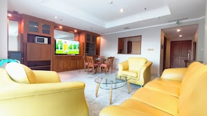 Sala de estar com sofás amarelos, uma mesa de centro de vidro, armários de madeira, televisão e um espaço de jantar com cadeiras e uma mesa.