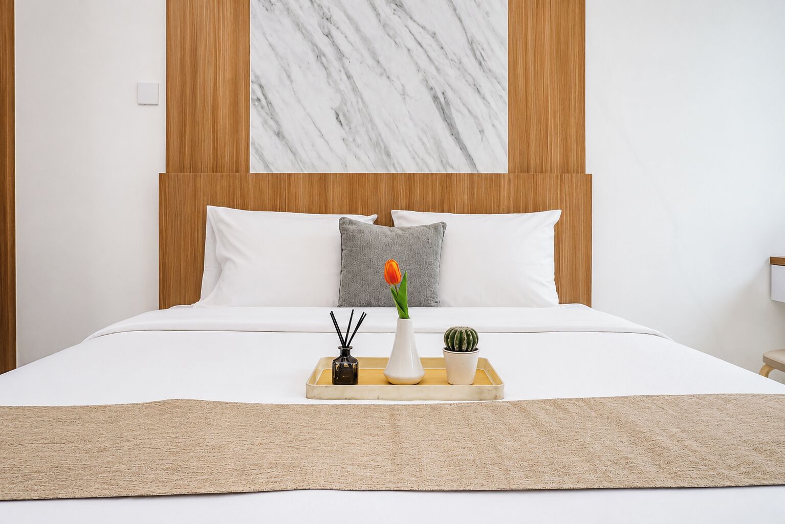 Una cama bien hecha con ropa de cama blanca, una almohada gris y una bandeja con un florero y una pequeña planta.
