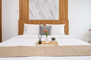 Una cama bien hecha con ropa de cama blanca, una almohada gris y una bandeja con un florero y una pequeña planta.