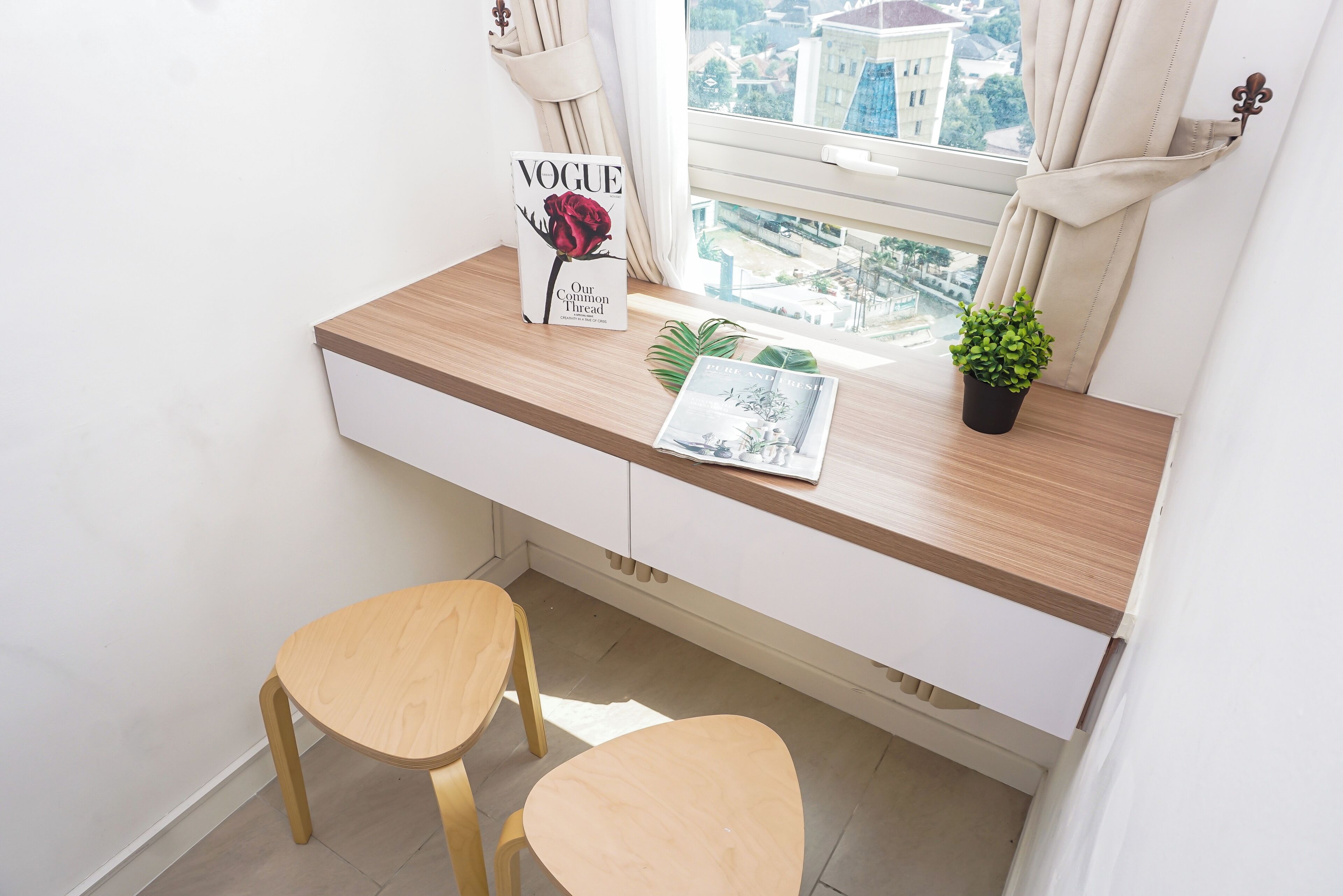 Una habitación pequeña y moderna con un escritorio, dos sillas de madera, un ventanal con vistas a edificios y una planta en maceta.