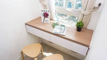 Una habitación pequeña y moderna con un escritorio, dos sillas de madera, un ventanal con vistas a edificios y una planta en maceta.