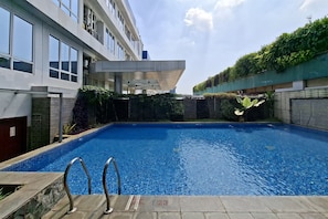 A swimming pool with a clear blue water surface, surrounded by a building with multiple windows and a covered area. - Warm Minimalist Studio At Apartment Harcosky Residence (Jakarta)