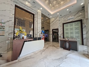 A hotel reception area with marble flooring, a reception desk, a decorative chandelier, and a wall-mounted air conditioning unit. - Warm Minimalist Studio At Apartment Harcosky Residence (Jakarta)