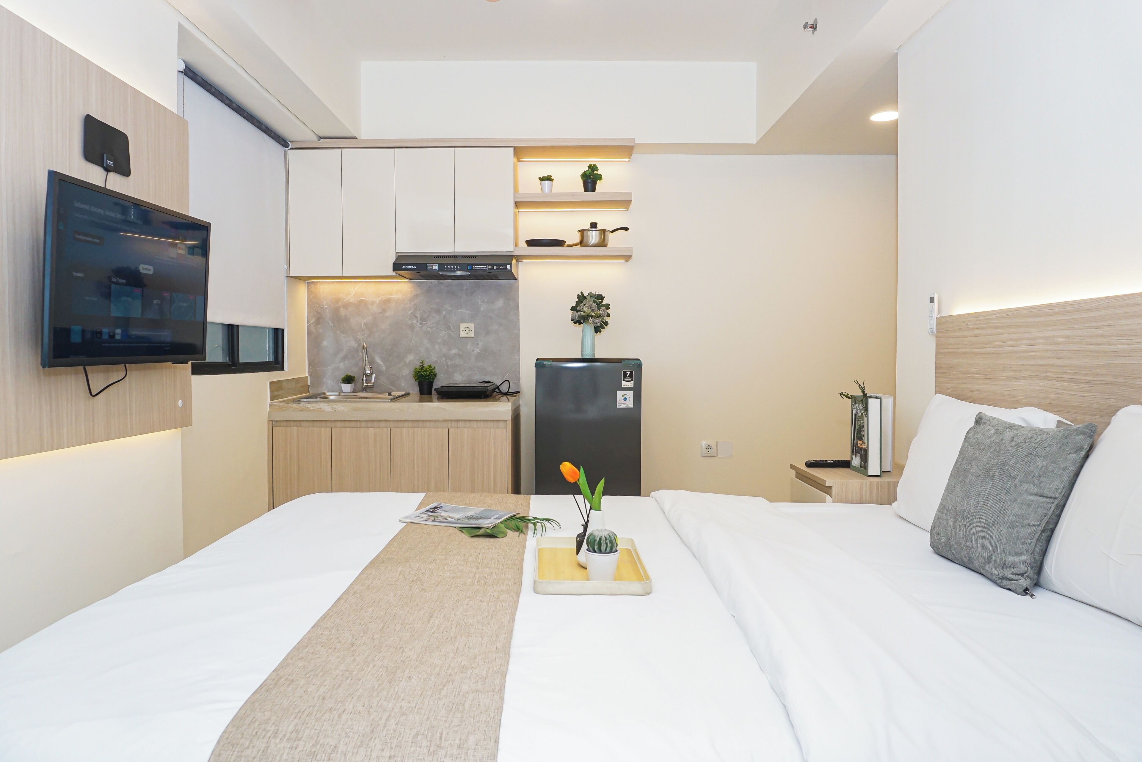 Habitación de hotel compacta con cama, mesita de noche, una pequeña kitchenette y un televisor montado en la pared.