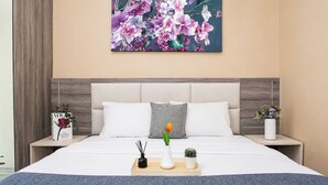 A modern bedroom with a large bed, a headboard, two bedside tables, a nightstand, a vase with flowers, a tray with a candle, and a framed picture on the wall. - Trendy 2Br At Apartment Vasanta Innopark (Cikarang)