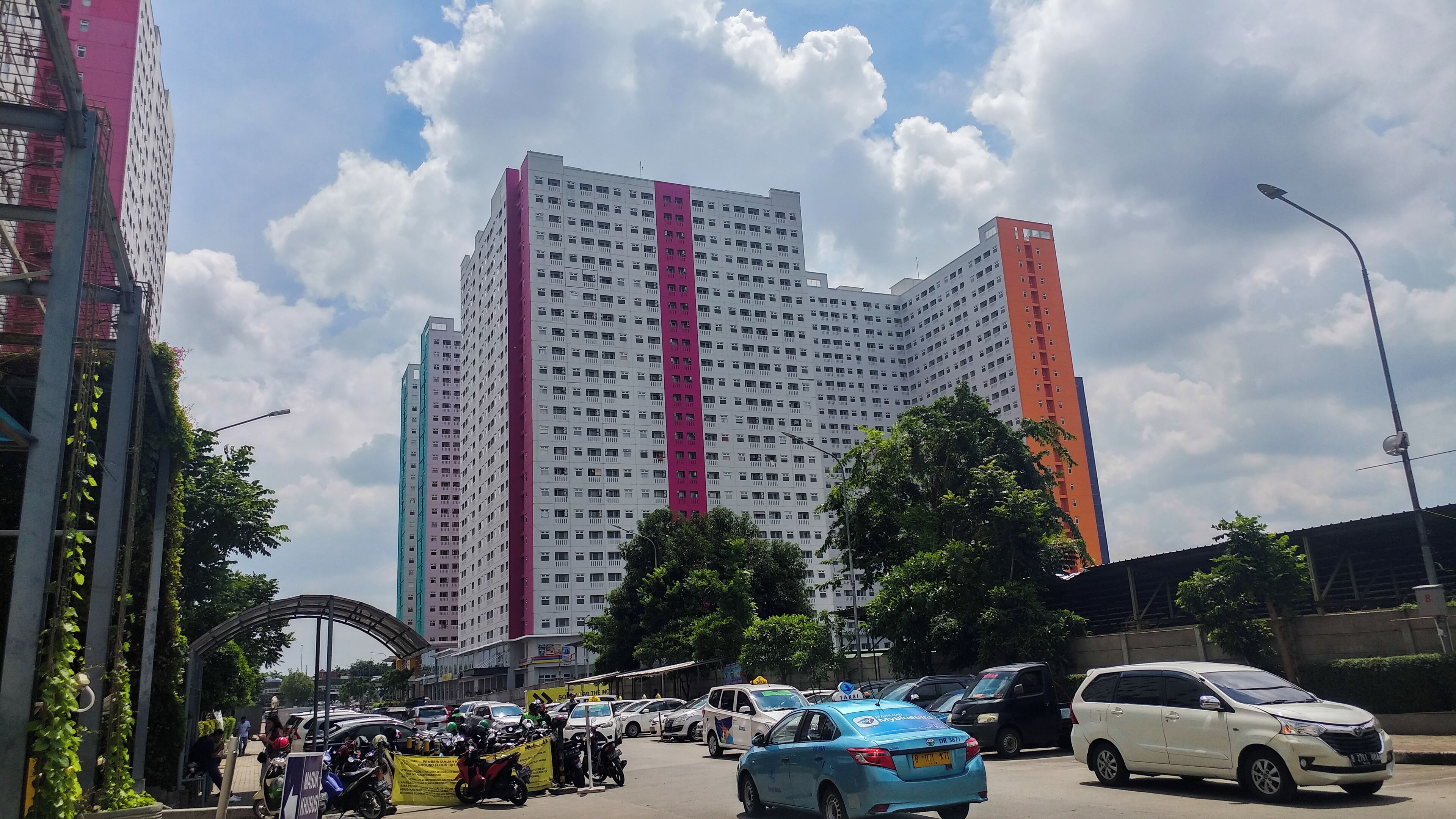 Un edificio de varios pisos con una llamativa banda rosa, rodeado de automóviles estacionados y vegetación.