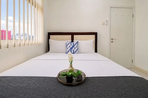 Quarto de hotel com cama, janela com persianas, porta e uma pequena mesa com uma planta.