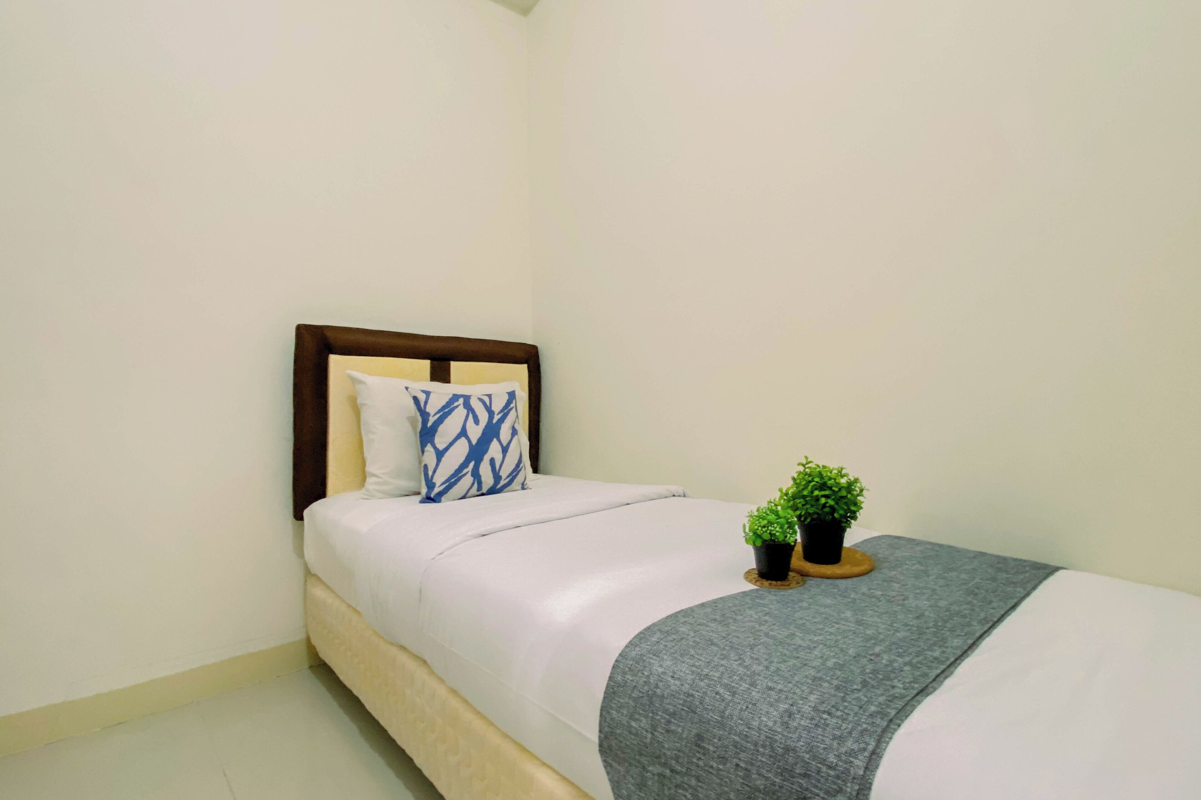 Uma cama de solteiro com cabeceira, uma mesinha de cabeceira com planta em vaso e um cobertor cinza.