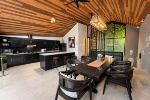 A modern kitchen with dark cabinetry, a central island, and a dining area with a long table and chairs. - Tropical 3BR Villa Coco Nusa Dua Bali (Nusa Dua)
