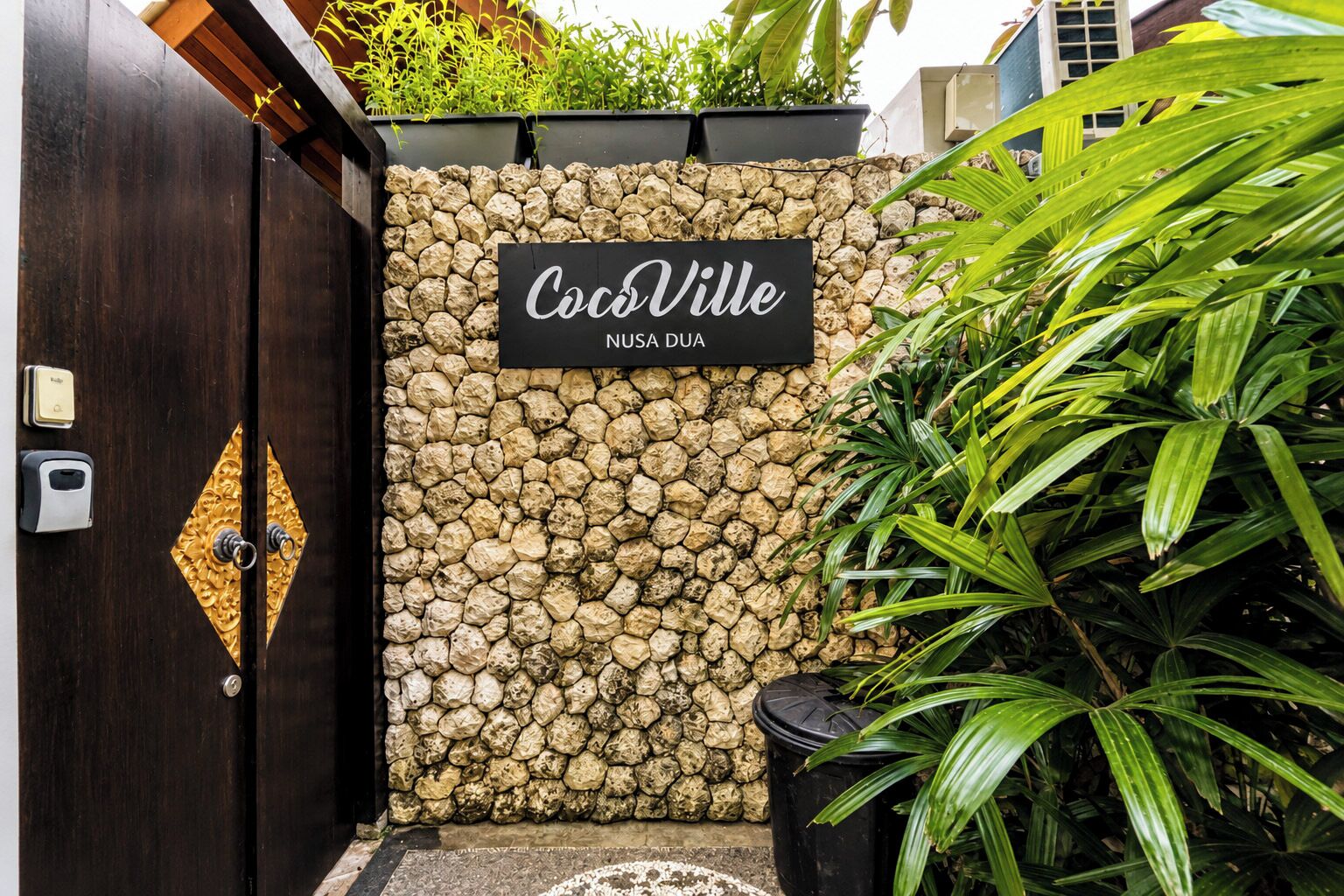 A stone wall with a sign reading 'CocoVille Nusa Dua' and a wooden door with a keyhole.