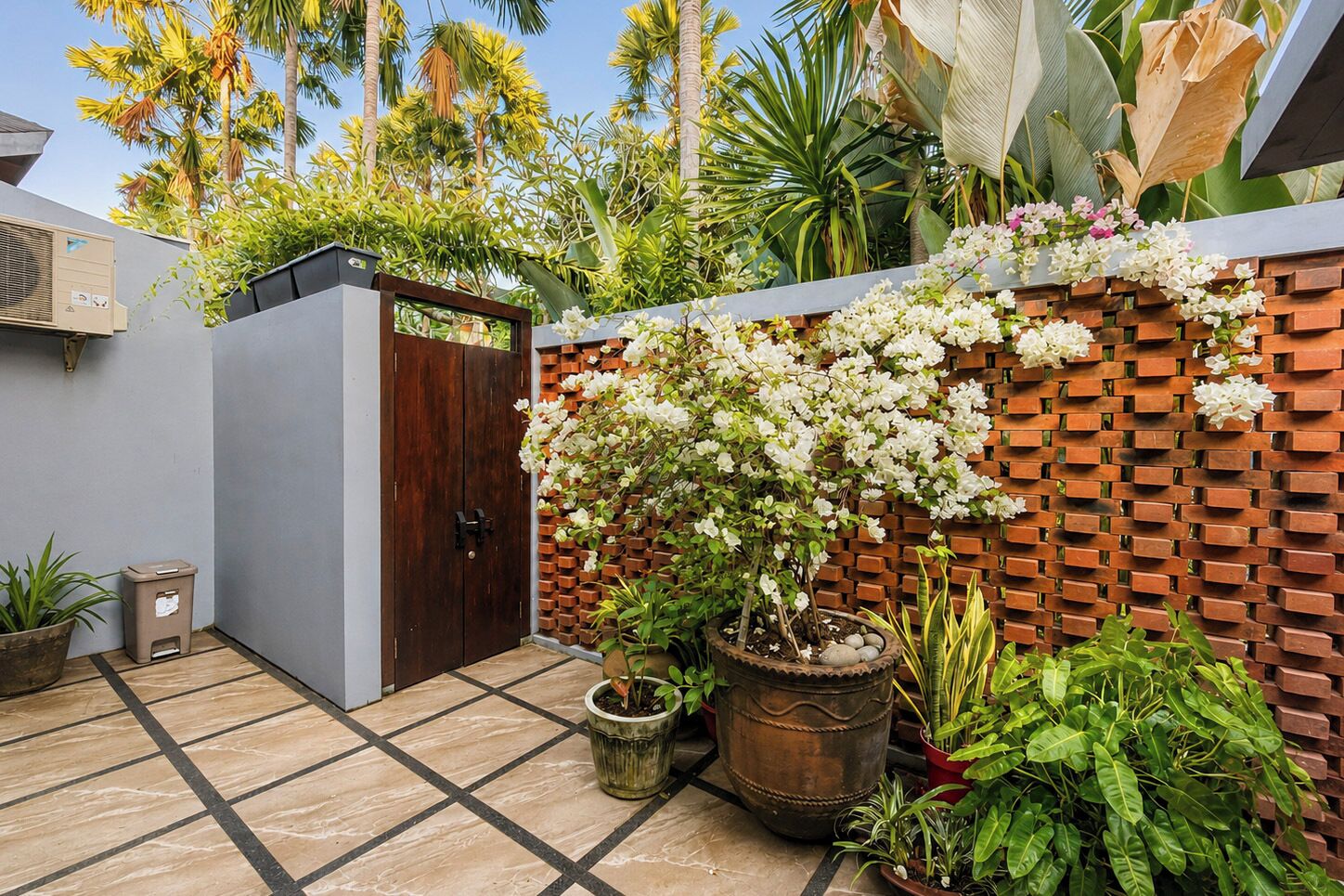 Une cour avec un sol carrelé, une porte en bois, des plantes en pot, un mur de briques orné de fleurs blanches et des plantes tropicales.