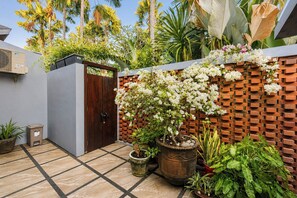 Un patio con piso de baldosas, una puerta de madera, plantas en macetas, un muro de ladrillo con flores blancas y plantas tropicales.