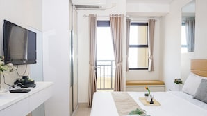 A hotel room with a bed, a desk with a computer and phone, a TV, and a balcony with curtains. - Convenient Studio At Apartment Transpark Cibubur (Depok)