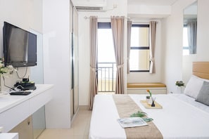 Habitación de hotel con cama, escritorio, computadora, teléfono, televisor y balcón con cortinas.
