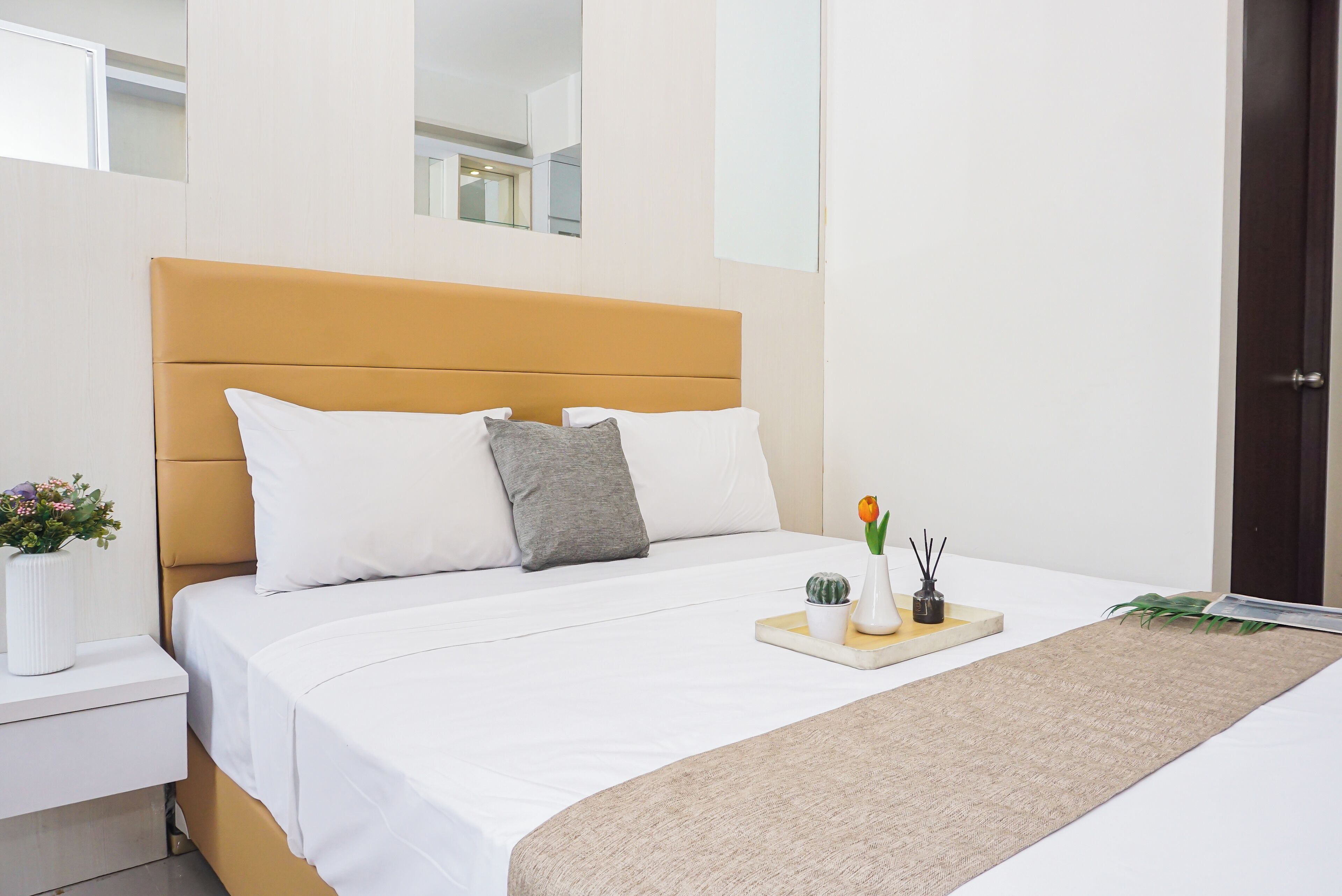 A modern bedroom with a bed, white bedding, a nightstand, a bedside table with a vase of flowers, and a tray with a plant and a candle.