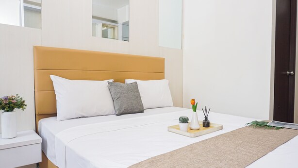 A modern bedroom with a bed, white bedding, a nightstand, a bedside table with a vase of flowers, and a tray with a plant and a candle. - Convenient Studio At Apartment Transpark Cibubur (Depok)