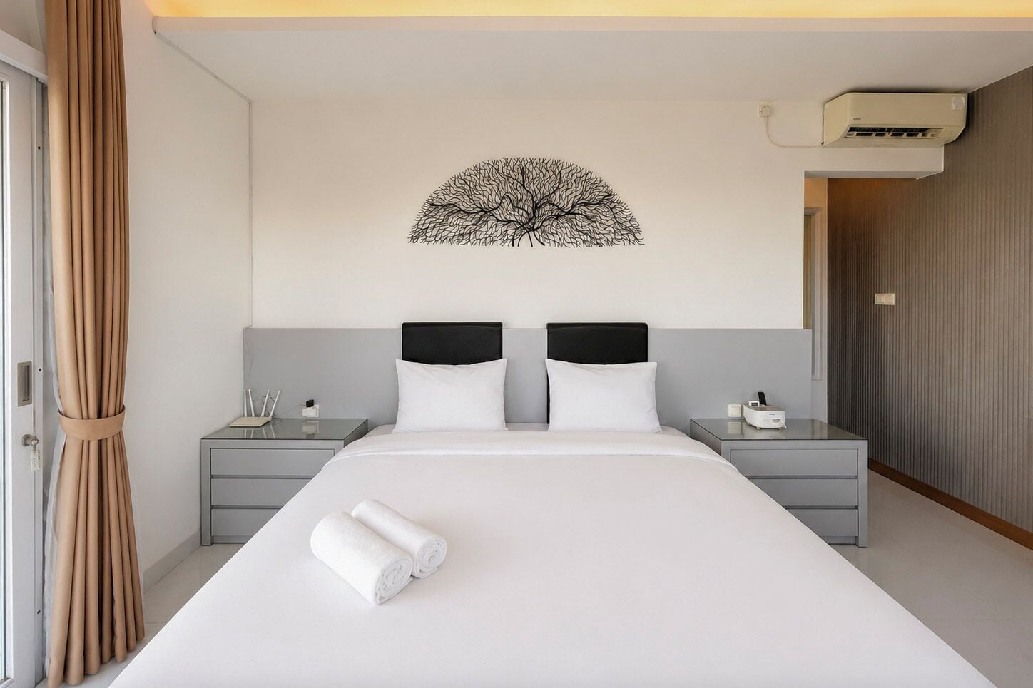 A hotel room with a large bed, two bedside tables, a wall-mounted air conditioning unit, and a curtain on the left.