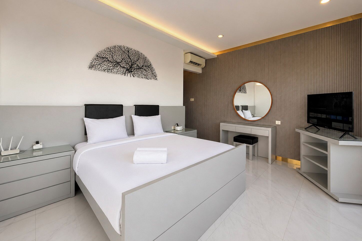 Ein modernes Hotelzimmer mit einem großen Bett, einem Nachttisch, einem Schreibtisch mit Stuhl, einem runden Spiegel, einem Flachbildfernseher und einer Klimaanlage.