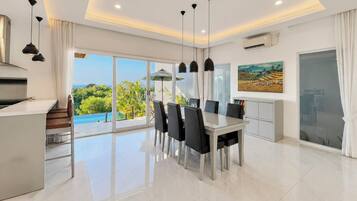 A modern dining area with a glass table and chairs, a kitchen with a built-in stove, and a large window offering a view of greenery and a pool.