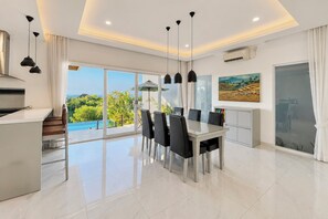 Ein moderner Essbereich mit Glastisch und Stühlen, eine Küche mit Einbauherd und ein großes Fenster mit Blick auf Grünflächen und einen Pool.