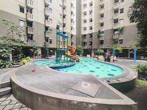 Ein Swimmingpool mit Rutsche und Spielplatz, umgeben von einer angelegten Grünfläche mit Bäumen und einem mehrstöckigen Gebäude im Hintergrund.