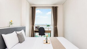 Een hotelkamer met een bed, een bureau, een stoel, een raam met gordijnen en uitzicht op de stad.