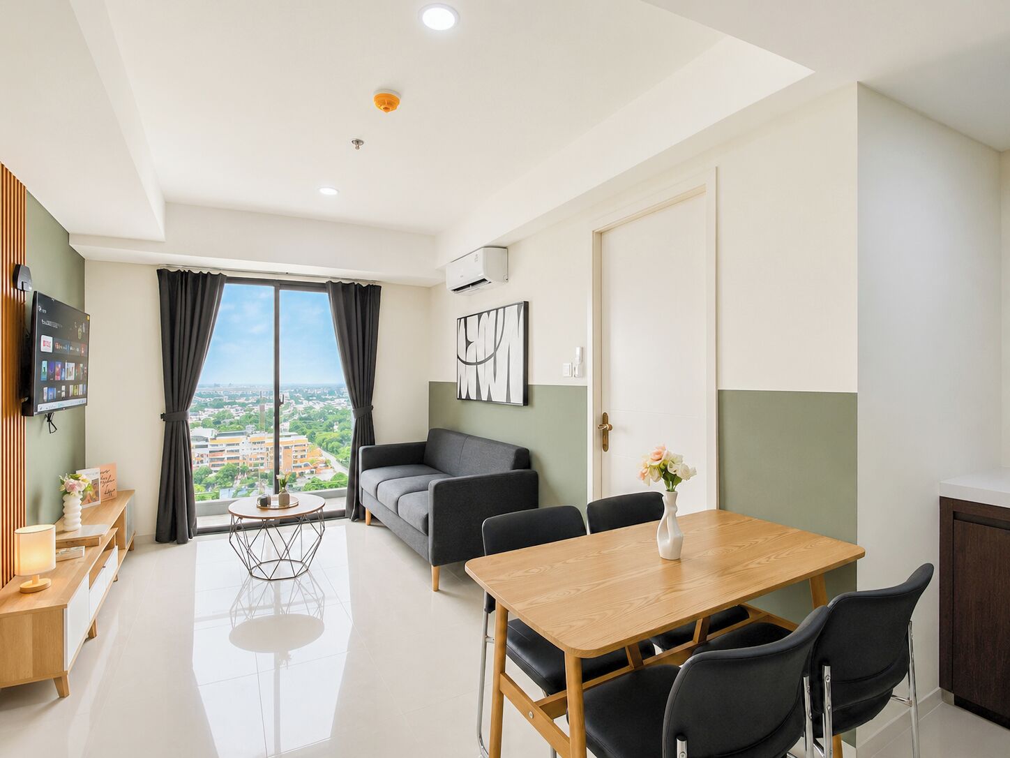 A modern living room with a sofa, a dining table with chairs, a TV mounted on the wall, and a large window with curtains.