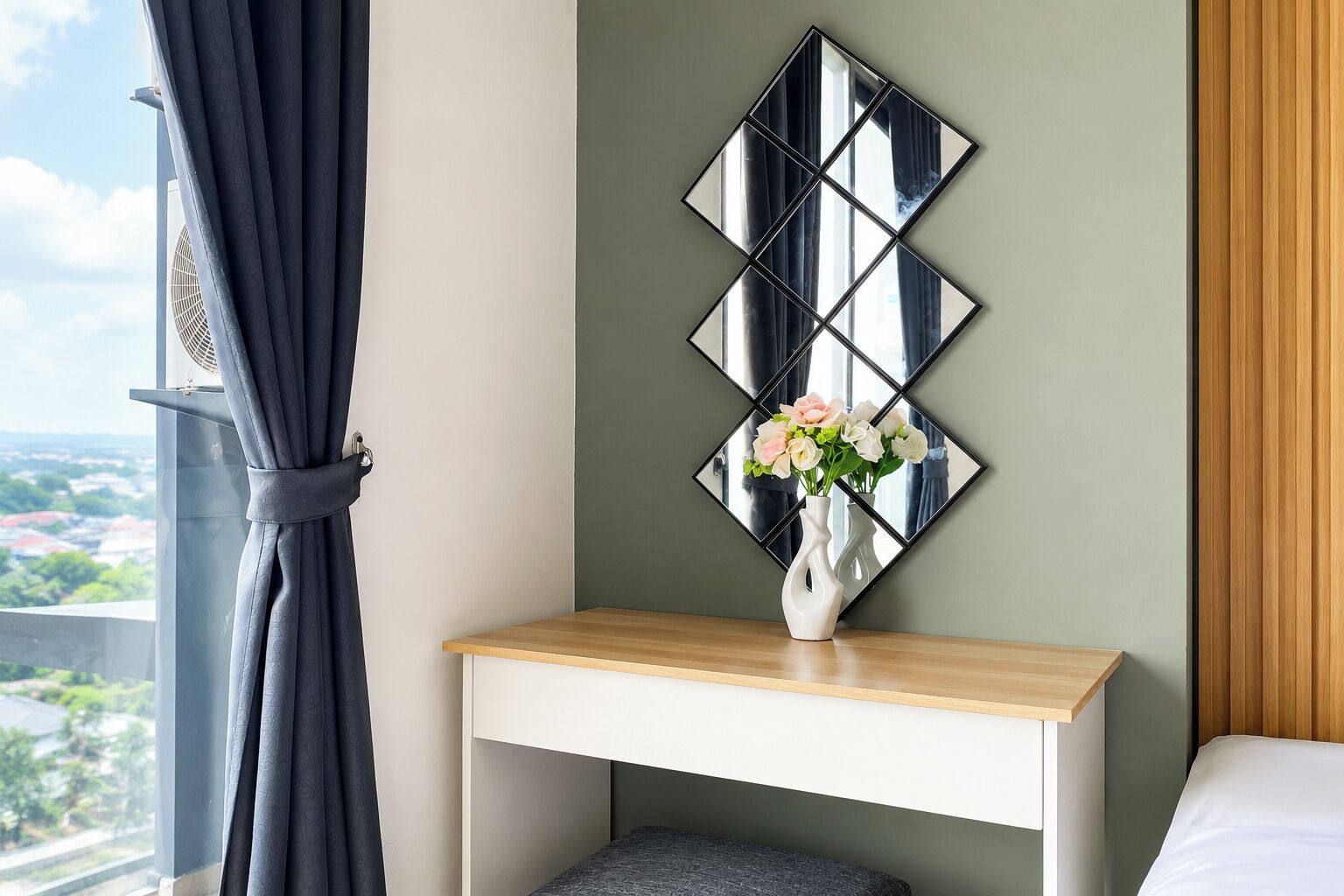A hotel room with a bed, a bedside table with a vase of flowers, a wall-mounted mirror, and a view of a cityscape through a window with curtains.
