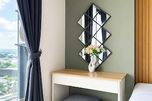 A hotel room with a bed, a bedside table with a vase of flowers, a wall-mounted mirror, and a view of a cityscape through a window with curtains. - Light Filled 3Br At Apartment Daan Mogot City (Jakarta)
