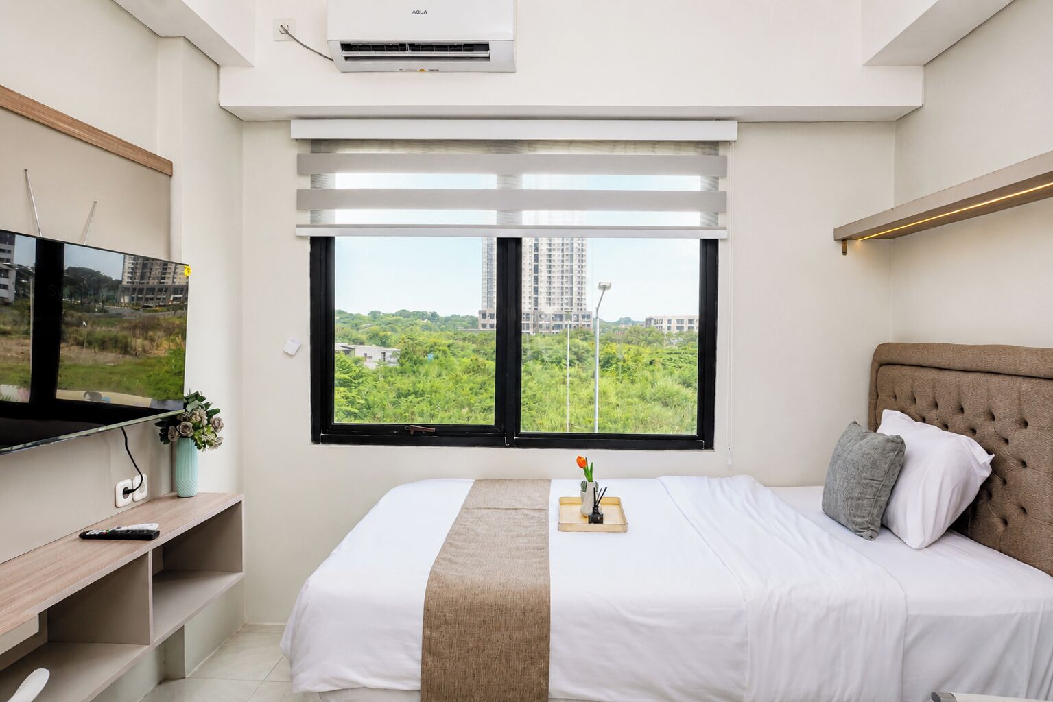 Ein modernes Schlafzimmer mit Bett, Fernseher und Fenster mit Blick auf Grünflächen und Gebäude.