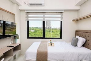 Ein modernes Schlafzimmer mit Bett, Fernseher und Fenster mit Blick auf Grünflächen und Gebäude.